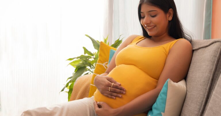 pregnant woman sitting on a couch while holding her belly with both of her hands.