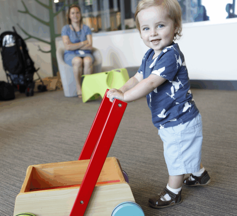 Toddler walking with the aid of a toy trolly. Mum watching in the background